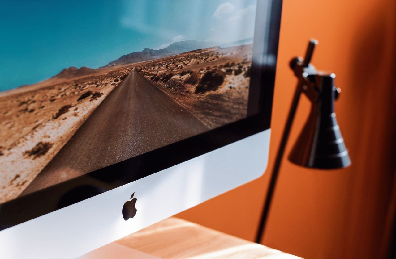 About A sleek modern workspace featuring an iMac displaying a desert road and a stylish desk lamp.